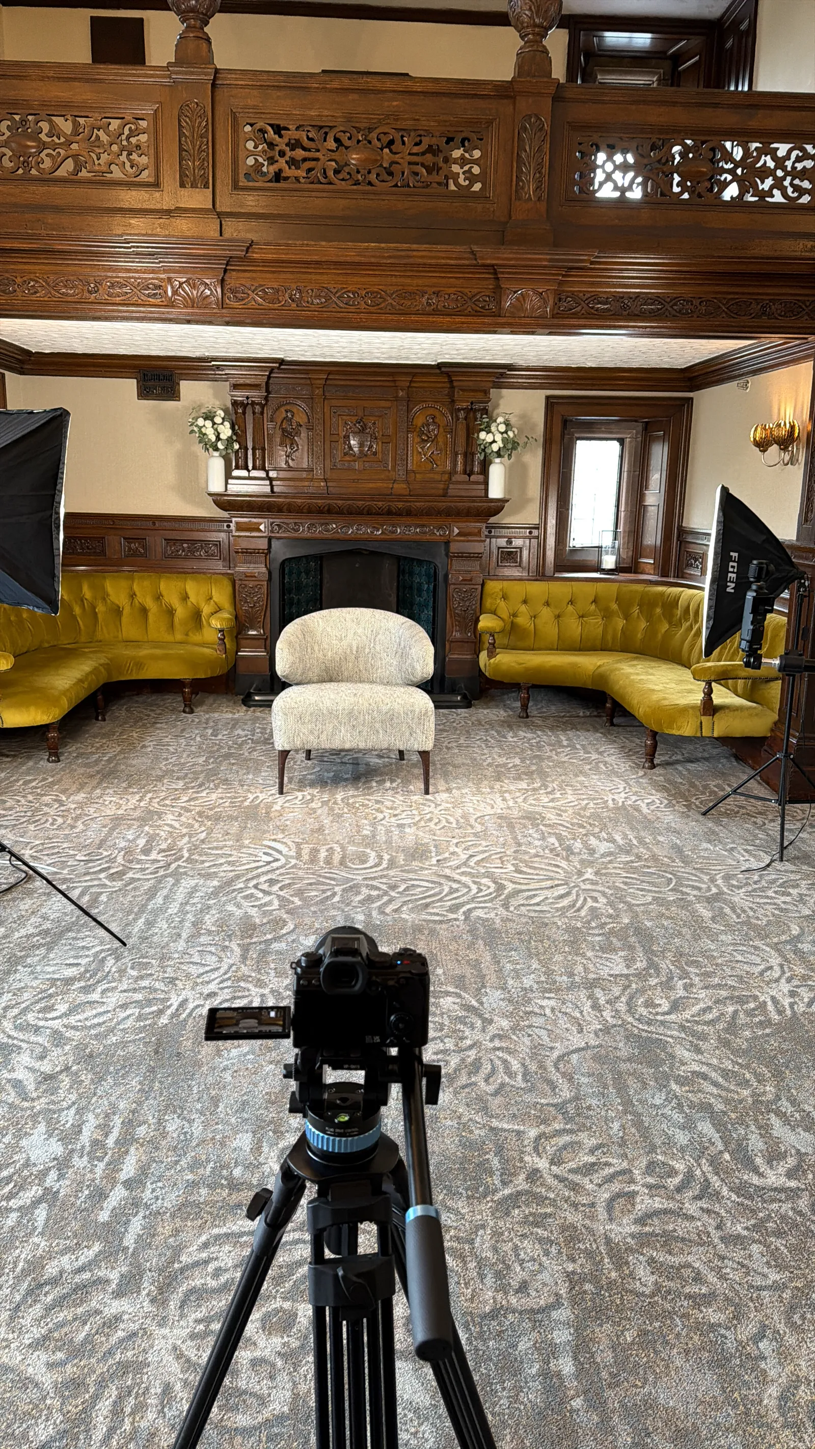 A camera on a tripod in a large wood-panelled room with lights and yellow sofas, set up for an interview