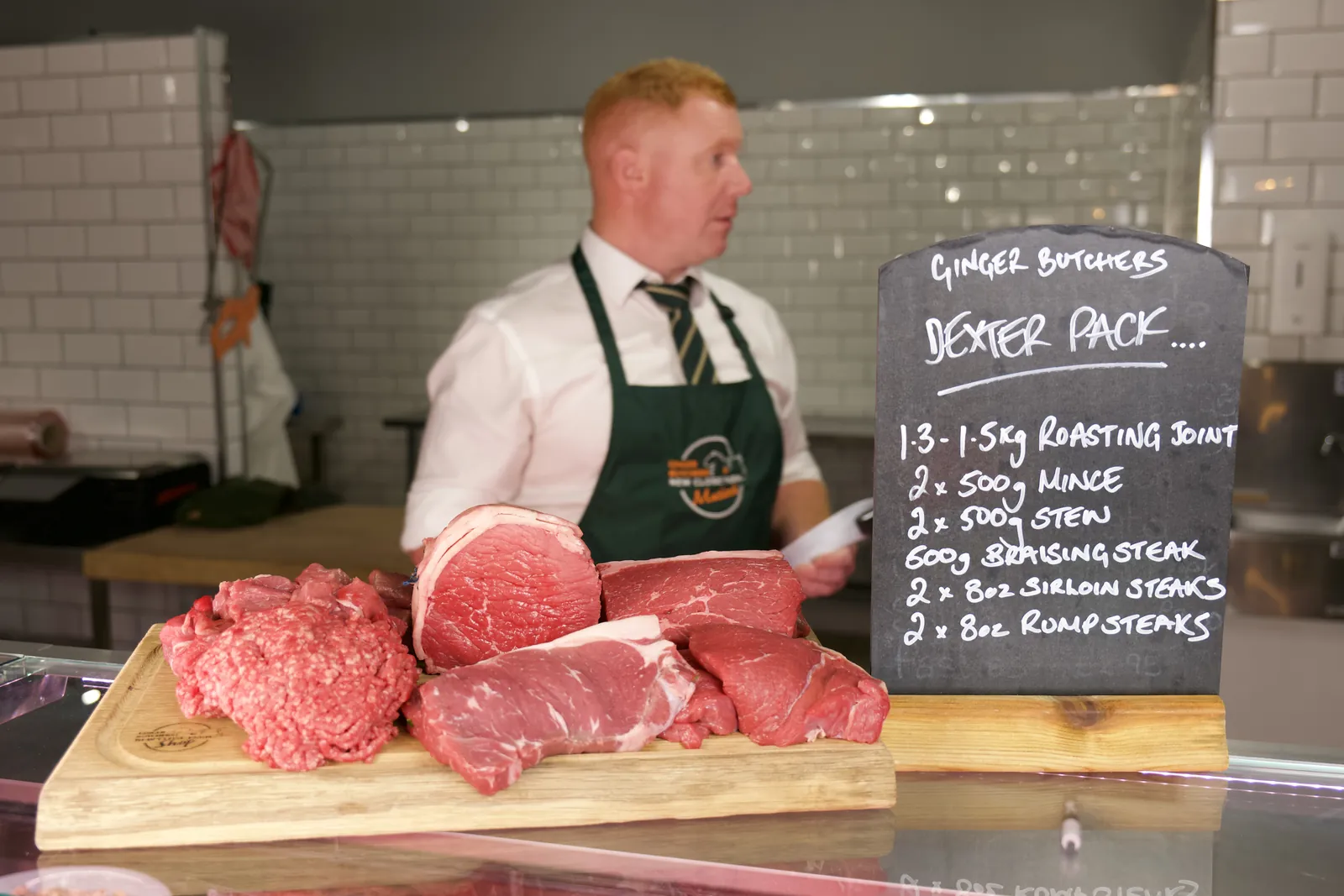 A butcher in a green Ginger Butchers apron stands behind a board of Dexter beef joints and mince next to a handwritten chalkboard listing the pack.