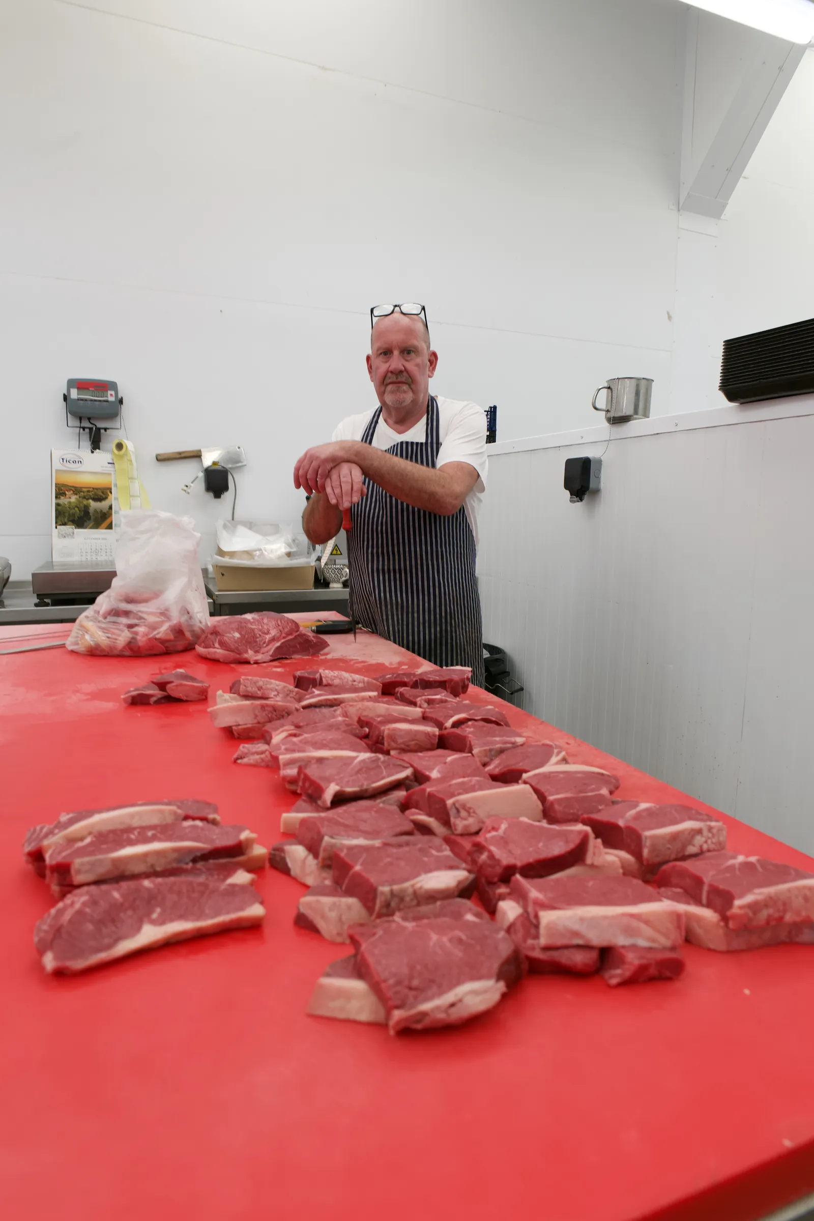 A butcher in a striped apron stands at the end of a red cutting board covered in freshly cut steaks.