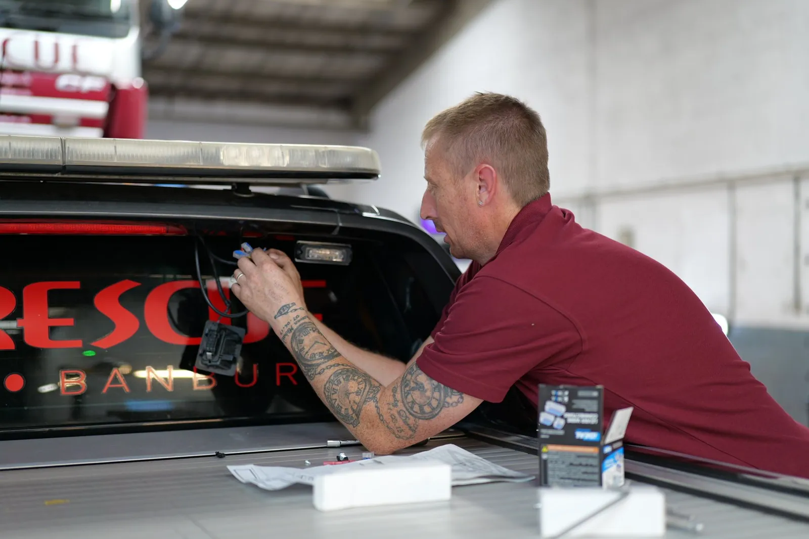 A tattooed technician in a maroon polo carefully applies red Isis Rescue lettering to the tail of a black recovery truck.