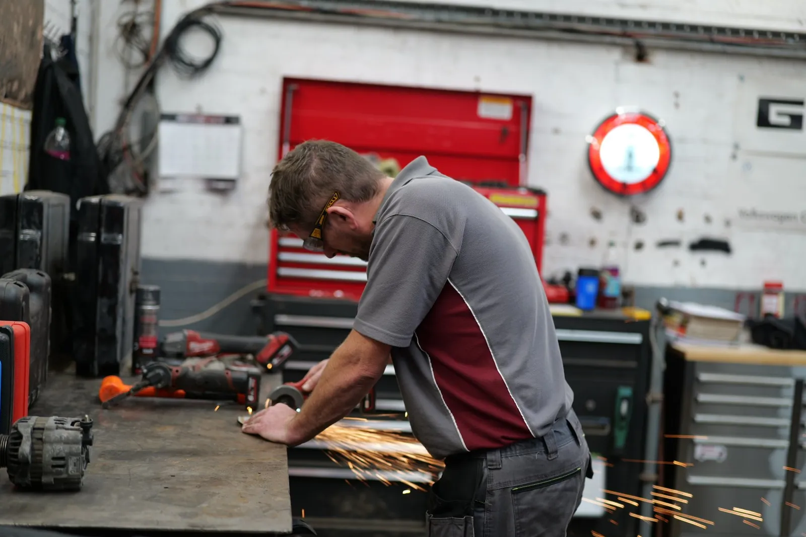 A technician in a maroon and grey uniform grinds metal at a workbench, throwing a shower of sparks, with a red tool chest behind him.