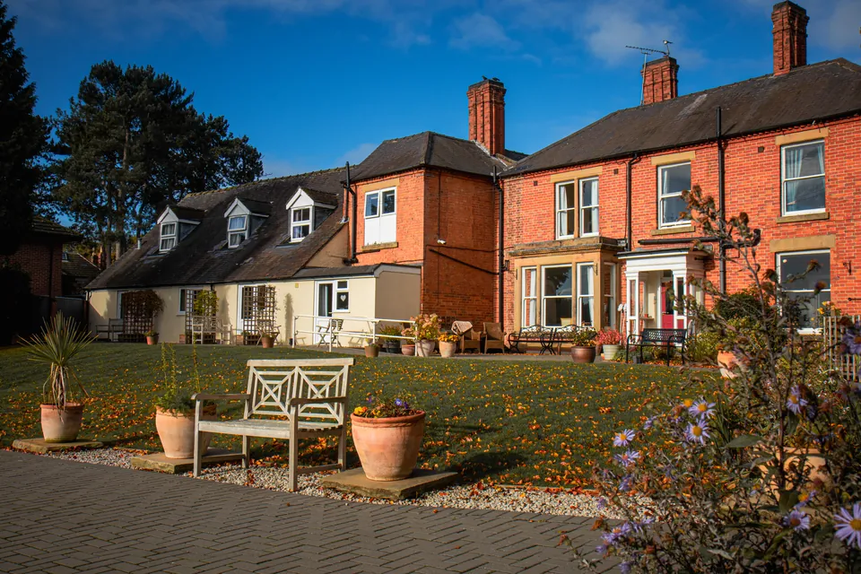 Ivy House care home's red brick buildings and gardens, with benches and planters in autumn sunshine