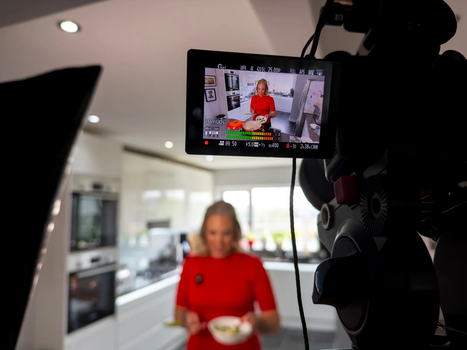 A presenter in a red top cooks at a kitchen island while a cinema camera, monitor and softbox light frame the shot in the foreground.