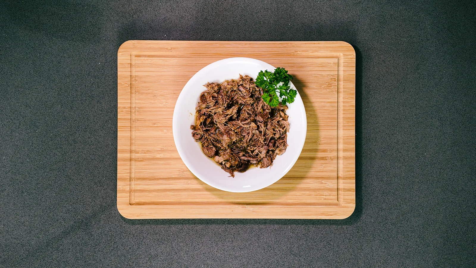 Slow-cooked garlic and herb lamb in a white bowl with parsley on a wooden board, from above