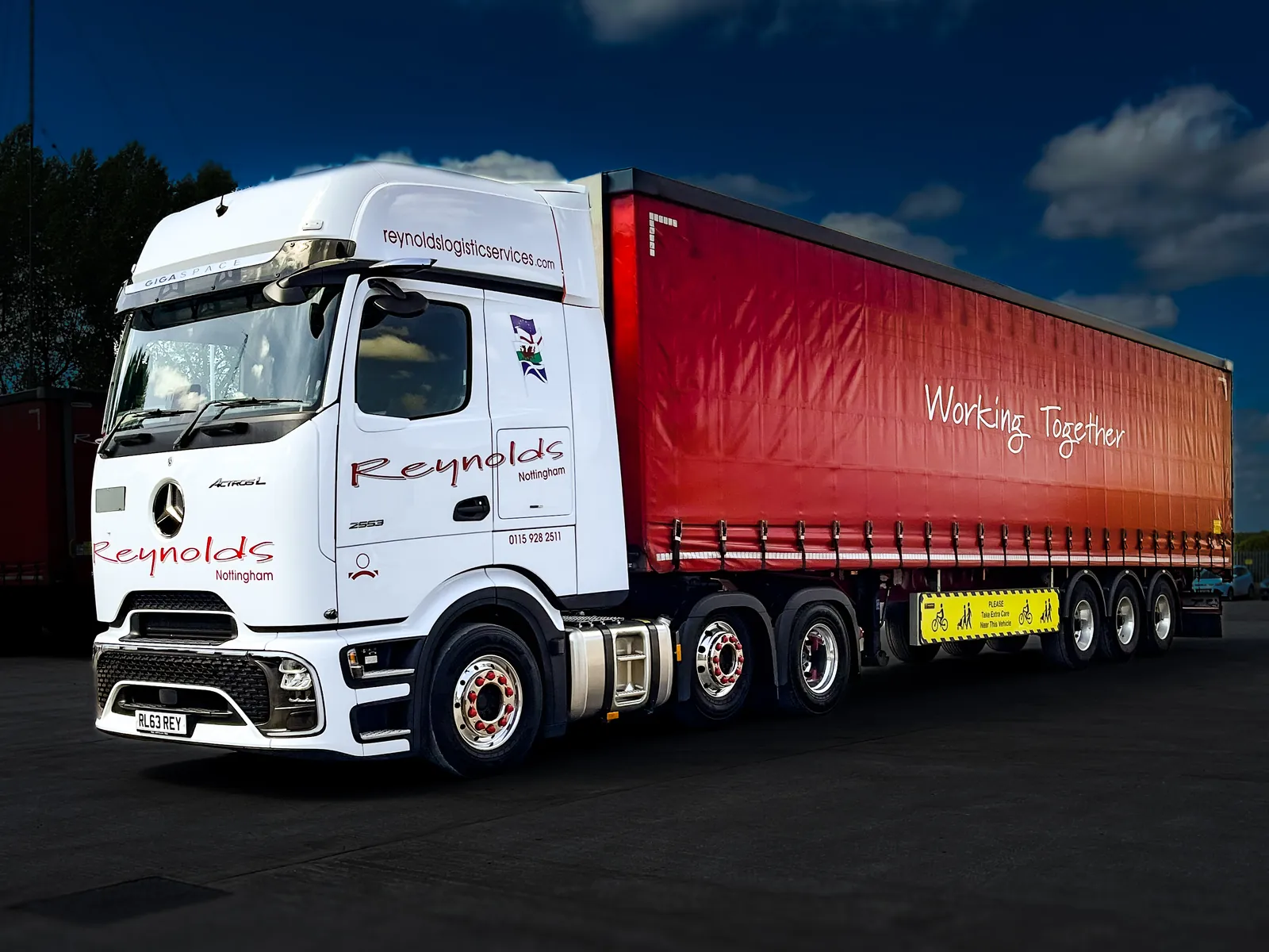 Low-angle shot of a white Mercedes Reynolds Logistics tractor unit pulling a red 'Working Together' curtainsider under a deep blue sky.