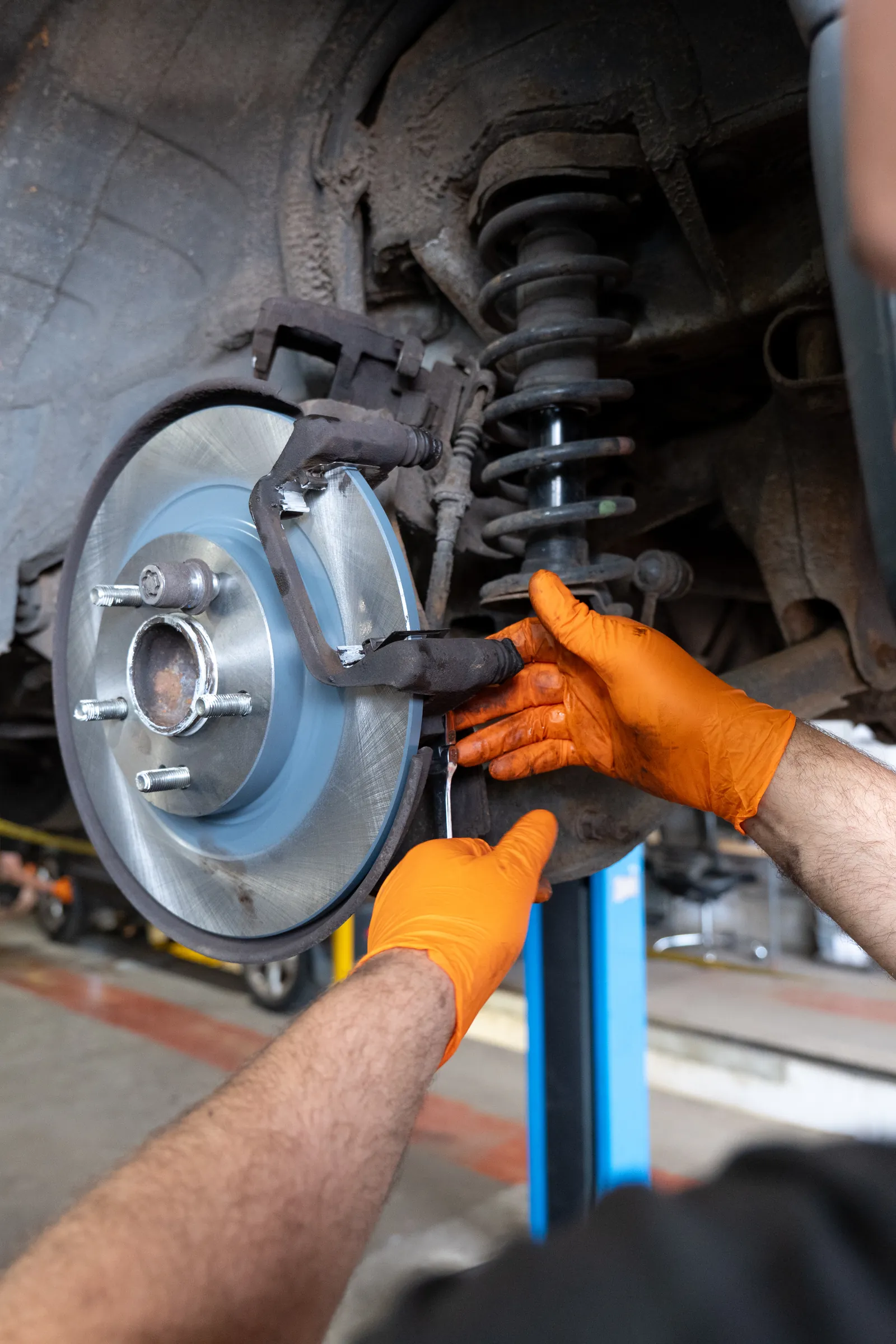 Close-up of a mechanic's hands in orange gloves fitting a brake caliper over a new disc beneath a car on a ramp.