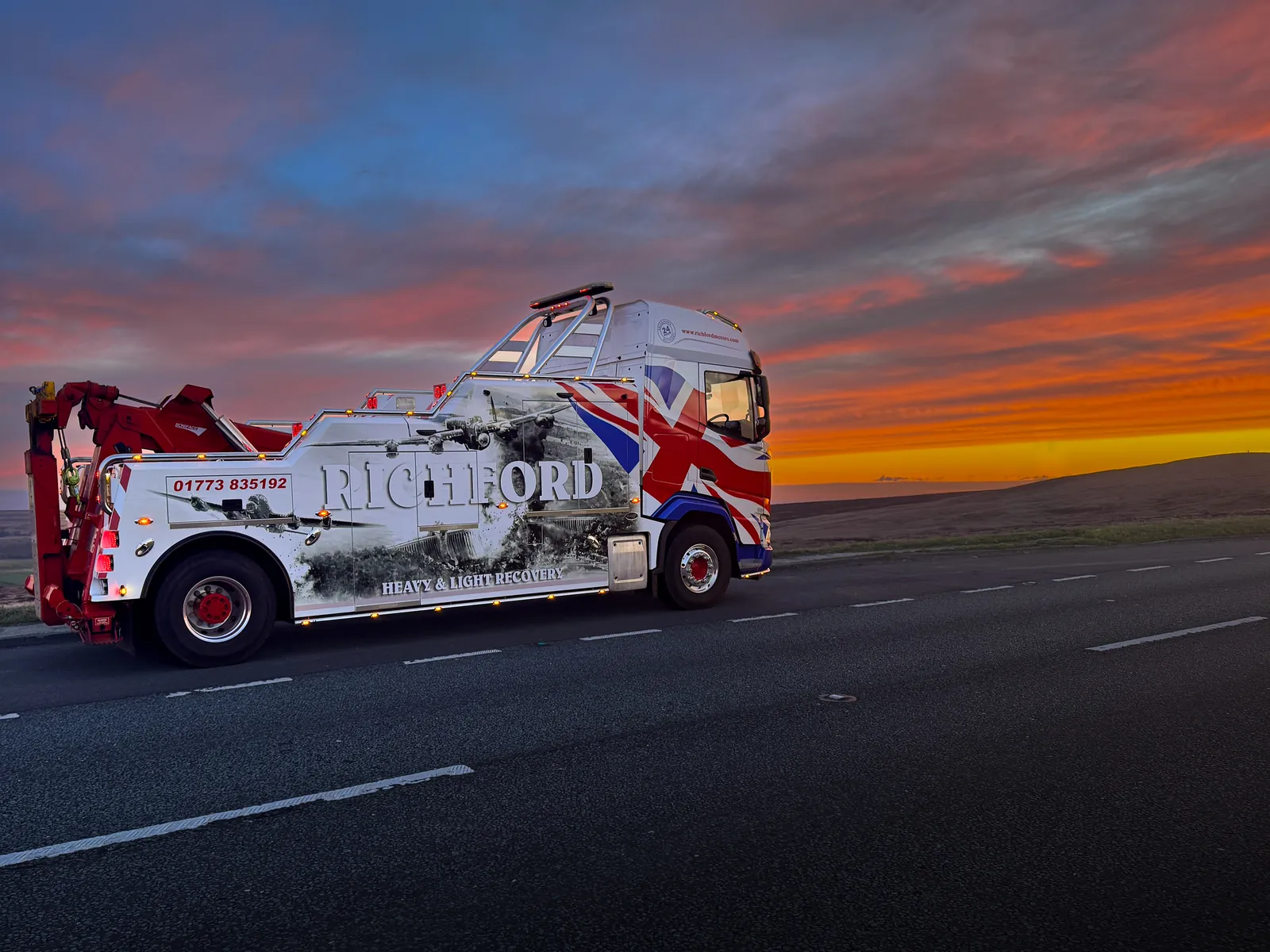 A white and red Richford heavy recovery truck is parked on a road under a vivid orange and purple sunset sky.