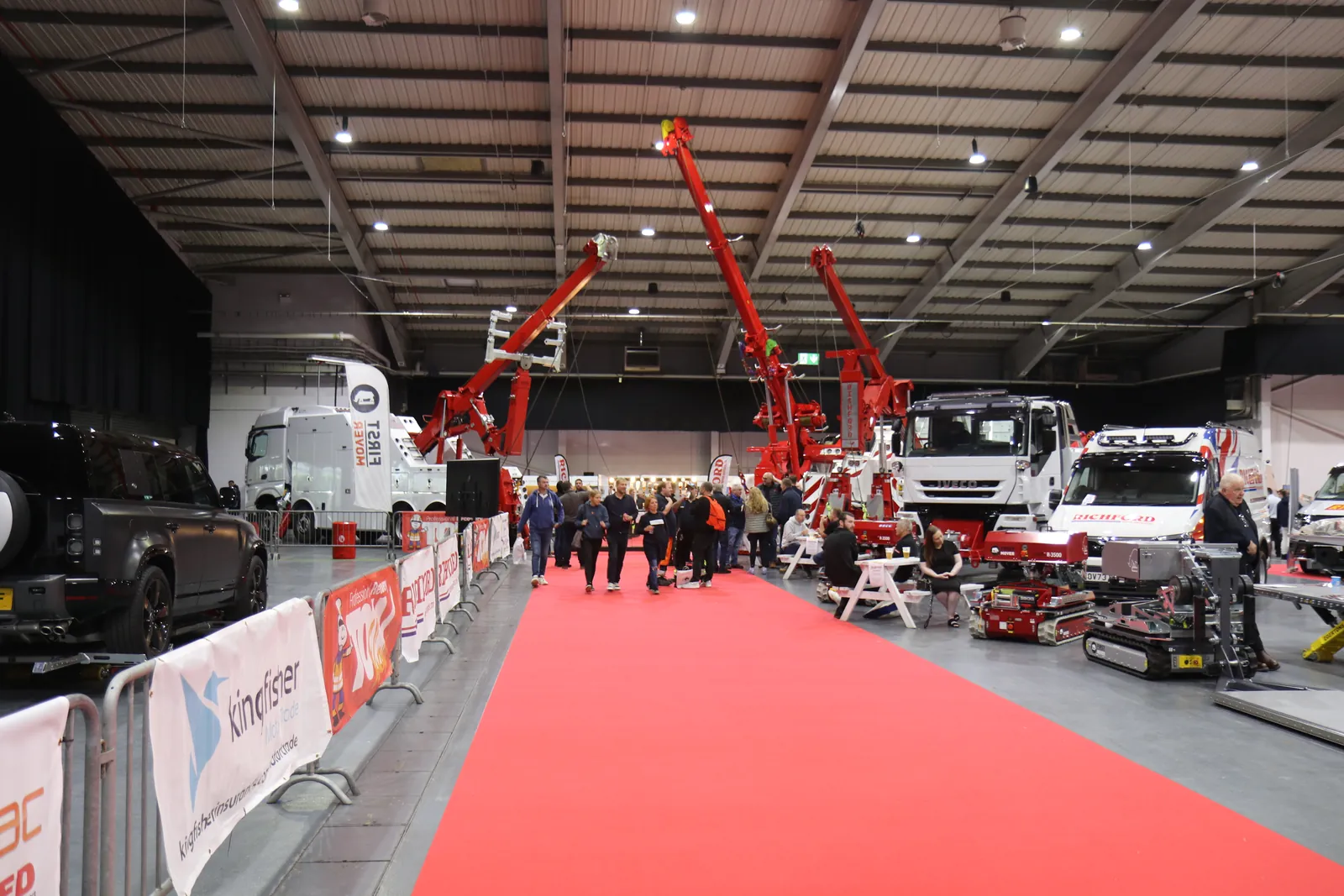A red carpet leads past barriers to recovery trucks with raised red cranes in a busy exhibition hall, visitors browsing the stands.