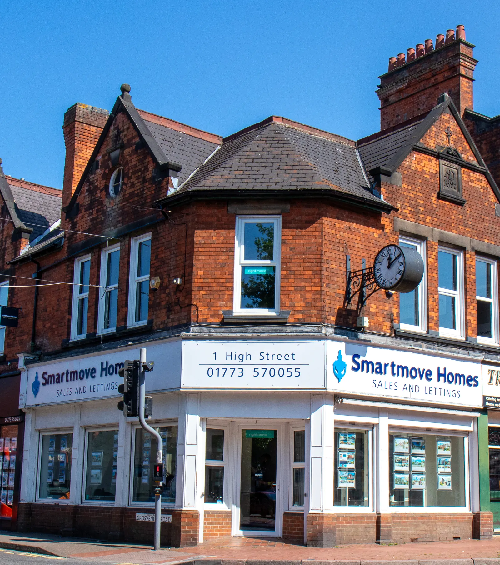 Upward view of the red-brick corner building housing Smartmove Homes, its white shopfront, clock and gabled roof against a clear blue sky.