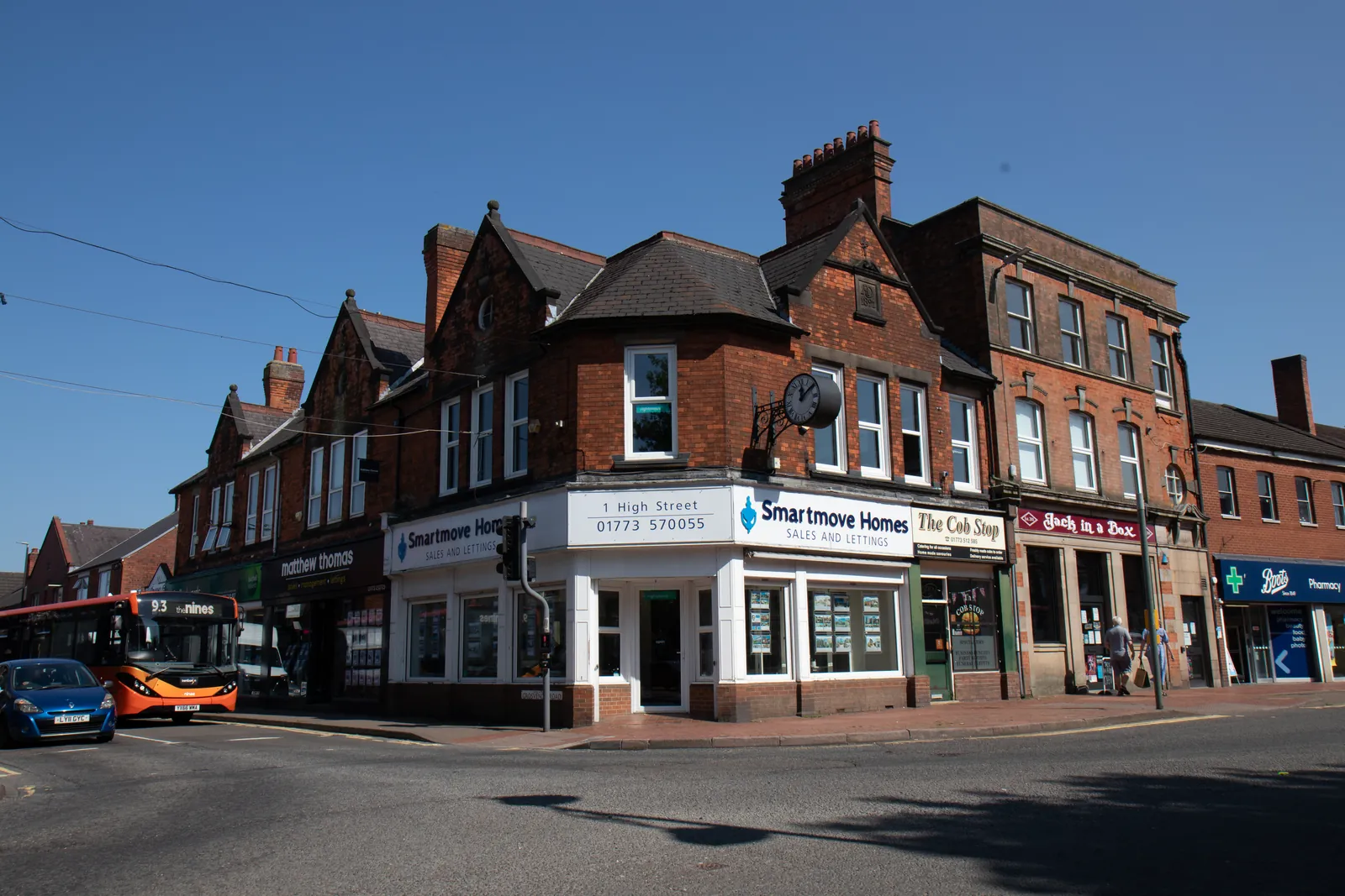 Wide street view of the Victorian red-brick terrace with the Smartmove Homes branch on the corner, a bus passing under a bright blue sky.
