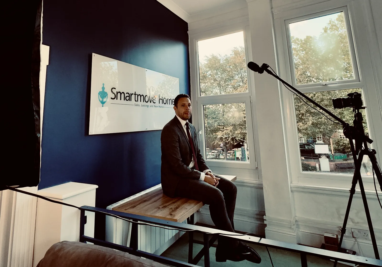 A suited estate agent sits on a bench in front of the Smartmove Homes sign while a camera on a tripod and a boom microphone record the interview.