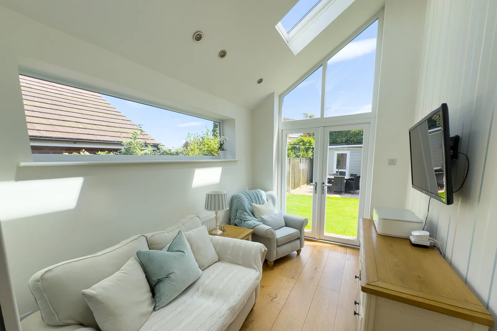 A sunlit garden room with a vaulted ceiling, skylight and tall glazed doors opening onto a lawn, a cream sofa in the foreground.
