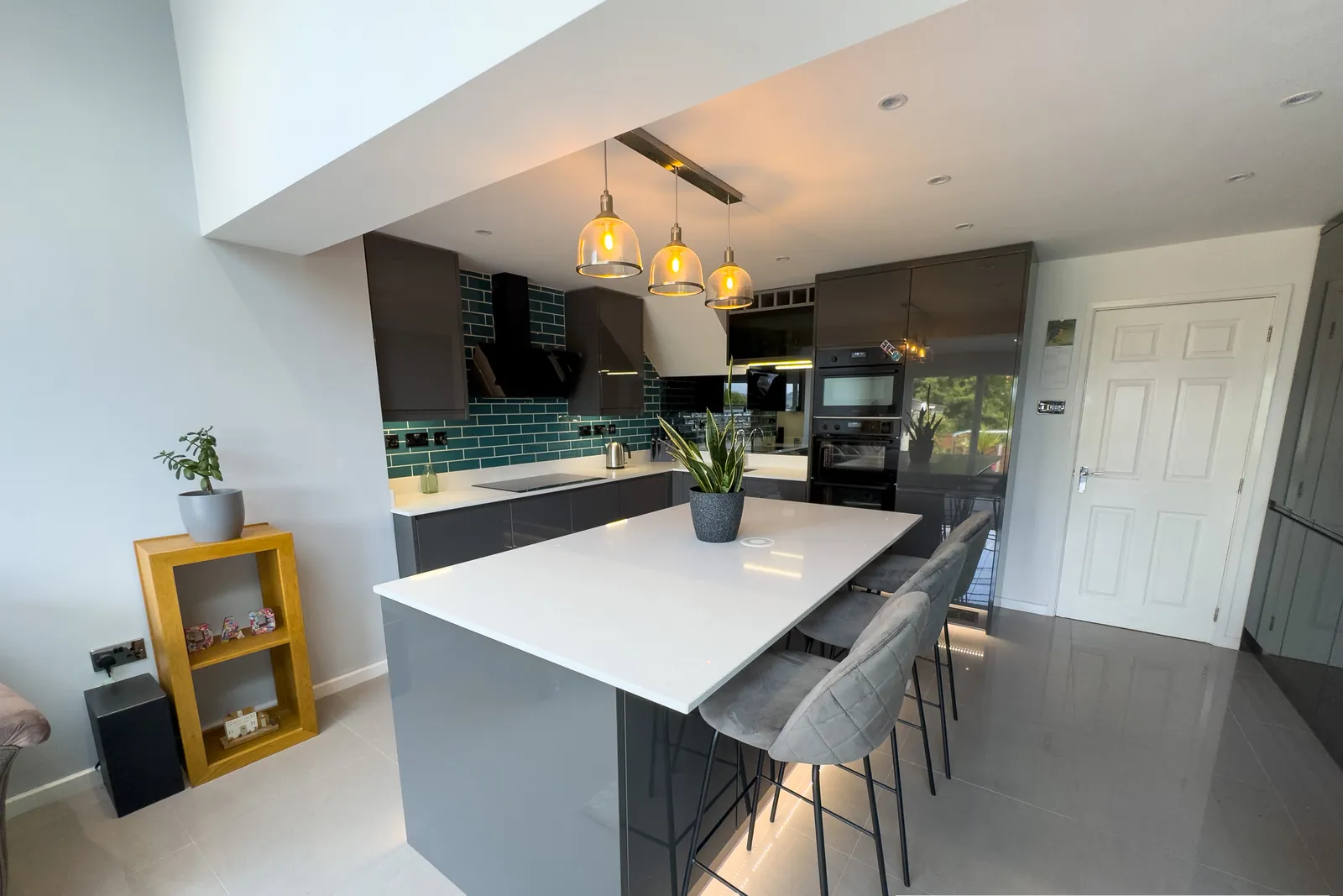 A modern grey kitchen with a white-topped island, pendant lights and bar stools, lit by a long window.