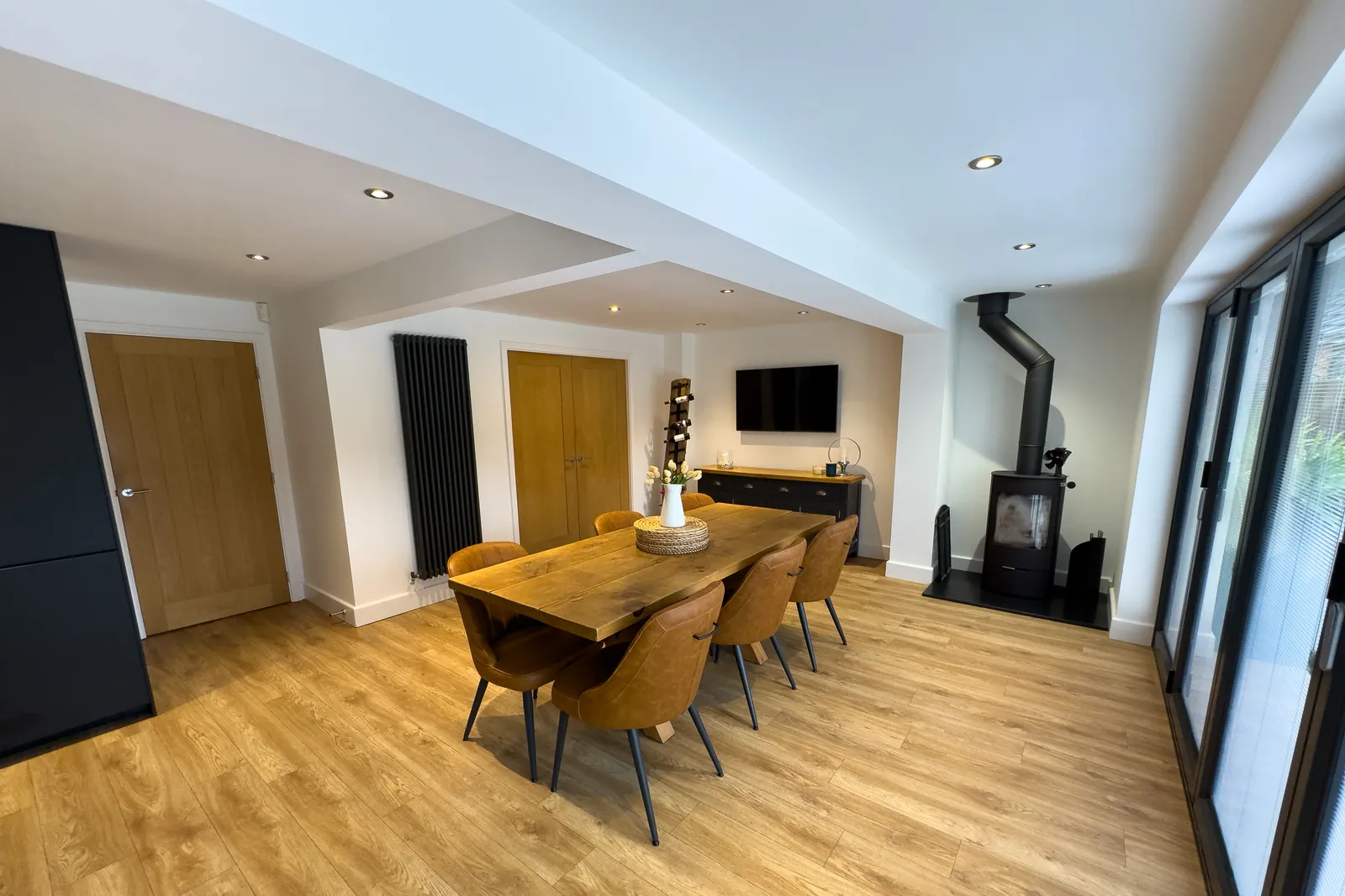 An open-plan room with oak flooring, a wooden dining table and chairs, and a log burner beneath a wall-mounted TV.
