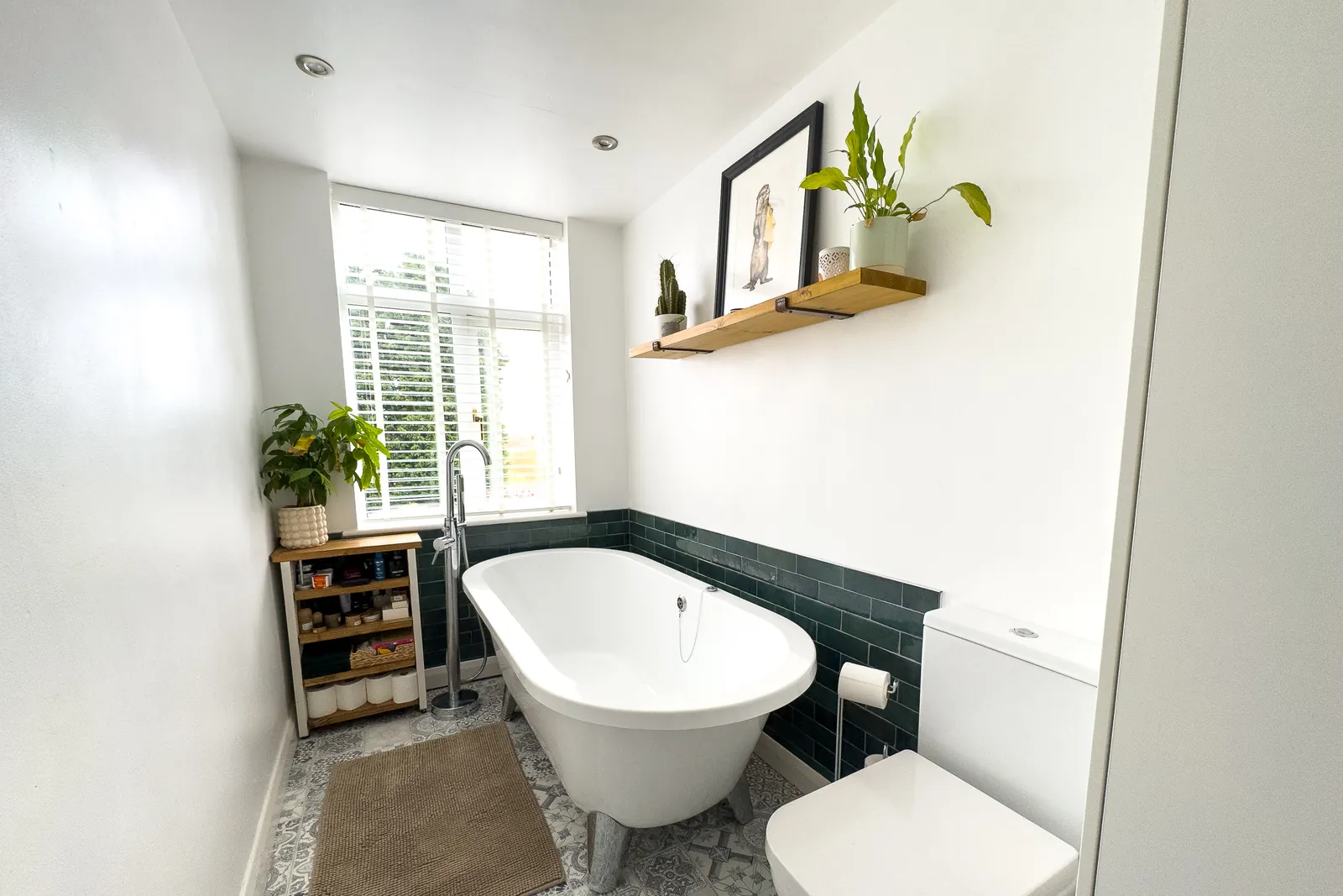 A white bathroom with a freestanding roll-top bath, patterned floor tiles, green tiled splashback and plants on a floating shelf.