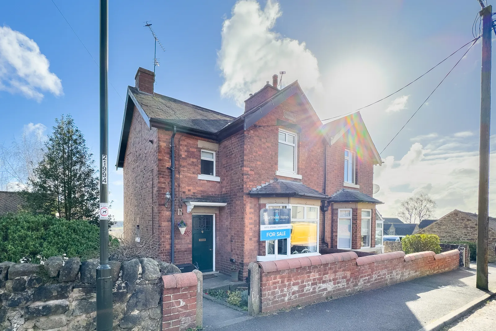 A red-brick Victorian semi-detached house with bay windows and a stone boundary wall, the sun flaring over its gable.