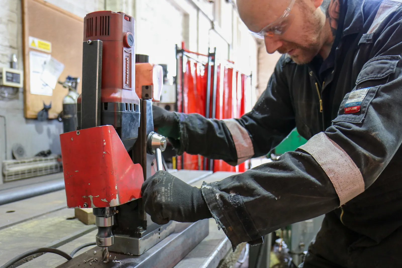 An engineer in black overalls and gloves operates a red magnetic drill on a steel section in the Sword Engineering workshop.