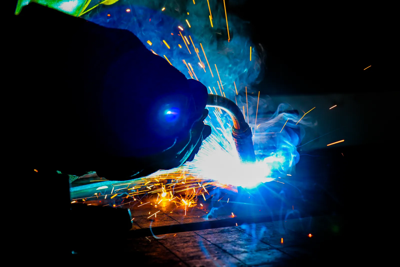 Close-up of a welder's gloved hand guiding a MIG torch, with blue arc light, smoke and orange sparks against a dark background.